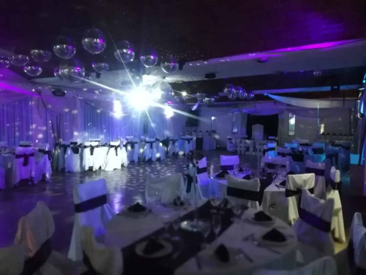 Pista del salón con esferas espejadas colgantes, luces violetas y mesas vestidas de blanco con moños negros