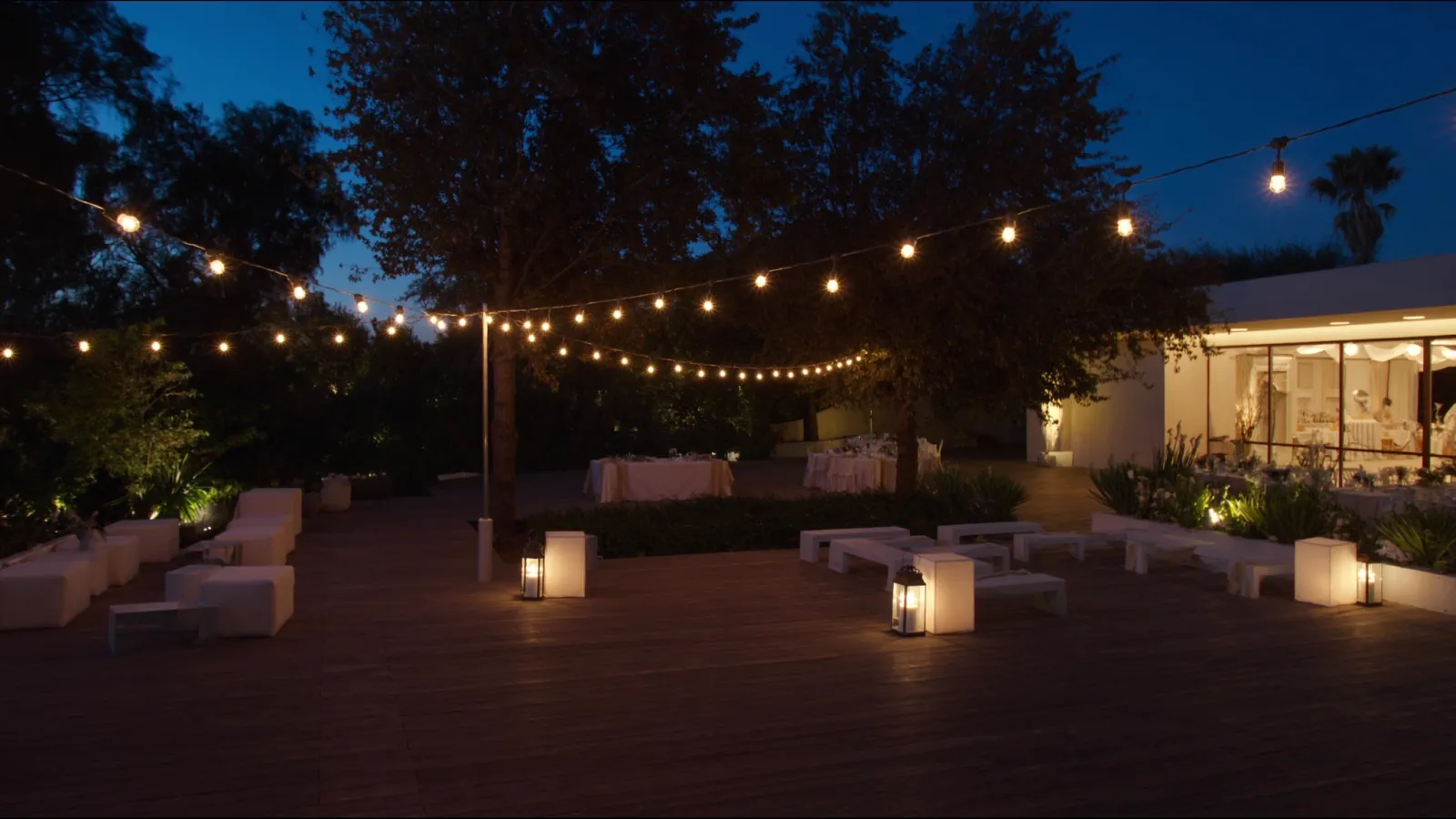Deck al aire libre con guirnaldas de luces cálidas, puffs blancos y faroles al atardecer
